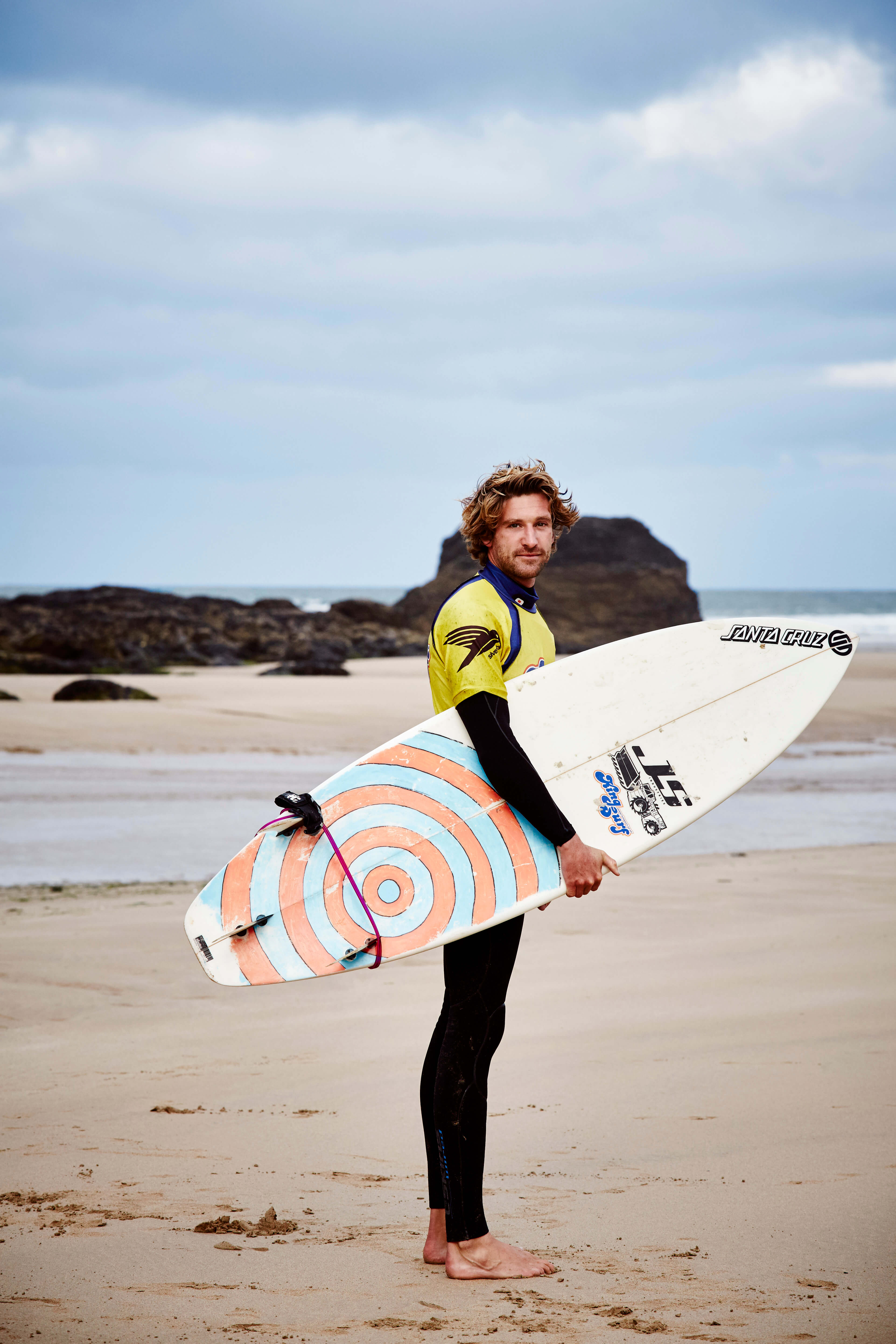 man standing on beach with a surfboard wearing a wetsuit and yellow rash vest