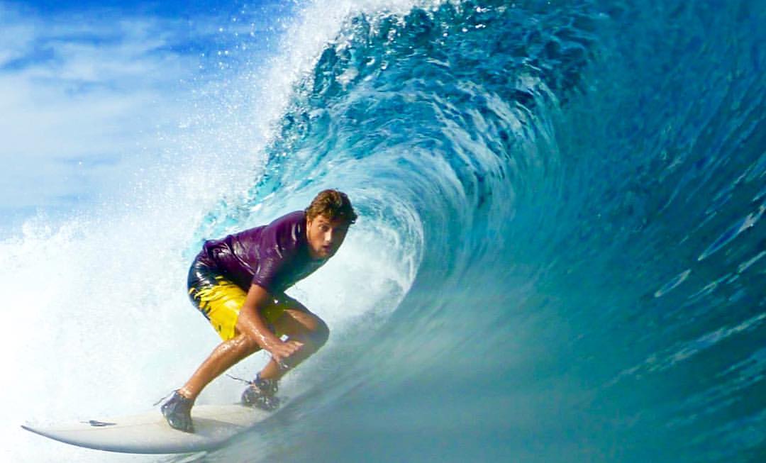 Man surfing barrel wave in purple rashvest and yellow boardshorts